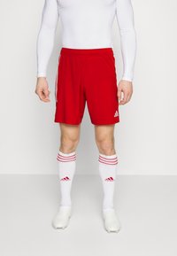 Rode sportieve shorts met een elastische tailleband en drie witte strepen aan elke zijkant, gedragen over witte sokken met rode strepen, gecombineerd met voetbalschoenen.