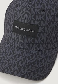 Kapa za bejzbol s uzorkom "MK" u tamnim tonovima i crna oznaka s natpisom "MICHAEL KORS" u sivim slovima.