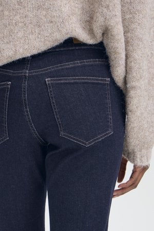 Gros plan d'une personne portant un jean bleu foncé avec des poches arrière visibles et un pull beige duveteux, main détendue sur le côté.