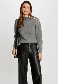 Pull en maille gris avec une texture côtelée, ajusté à la taille, avec des boutons décoratifs sur l'épaule gauche ; associé à un pantalon large en cuir noir.