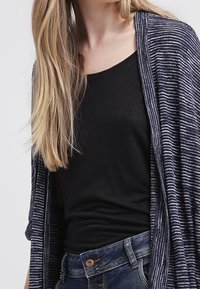 Cardigan blu scuro con un motivo a righe testurizzato, indossato sopra un top nero attillato. Abbinato a pantaloni di denim blu con chiusura a bottone.