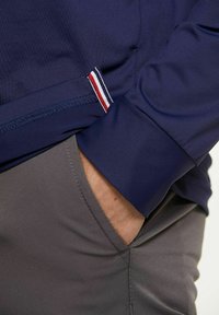 Marineblaue Jacke mit Kontrastdetails in Rot und Weiß an den Ärmeln, kombiniert mit grauen Hosen. Glatter Stoff, taillierte Passform.