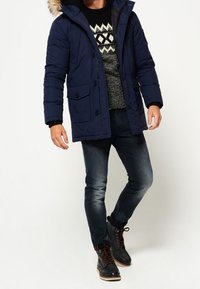 Chaqueta acolchada azul marino con ribete de piel sintética en la capucha, múltiples bolsillos delanteros, combinada con un suéter estampado, jeans oscuros y botas marrones.