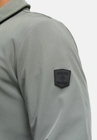 Hellgrüne Jacke mit samtiger Textur und einem schwarzen Stoffpatch am Ärmel, geprägt mit "INDICODE" und einem Logo.