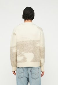 Kremasti džemper s mekom teksturom, s apstraktnim uzorkom u svijetlo bež boji. Rebrasti manšeti i rub, opušteni kroj.