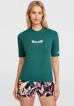 Femme portant un rashguard à manches courtes bleu sarcelle avec un logo blanc et un short de bain à motifs tropicaux colorés, debout devant un fond uni.