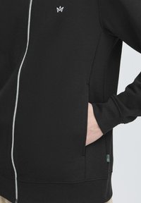 Hoodie noir avec fermeture éclair blanche, poches latérales et un subtil détail de logo. Texture de matériau douce et design ajusté, avec des poignets côtelés.