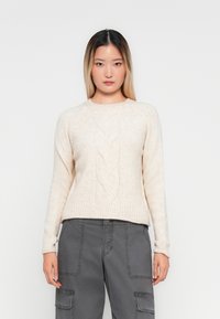 Donna con lunghi capelli scuri tendenti al biondo, che indossa un maglione a trecce color crema e pantaloni cargo grigi, in piedi di fronte a uno sfondo bianco semplice.