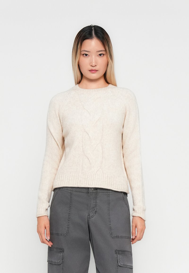 Donna con lunghi capelli scuri tendenti al biondo, che indossa un maglione a trecce color crema e pantaloni cargo grigi, in piedi di fronte a uno sfondo bianco semplice.