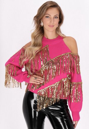 Roze gebreide sweater met gouden ceintuursfranje, cold shoulder ontwerp, lange mouwen; gedragen met glanzende zwarte broeken.