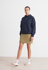 Námornícka mikina s kapucňou a šnúrkou, v kombinácii s khaki sukňou s predným rozparkom; modelka má na sebe biele tenisky s hnedými akcentmi.