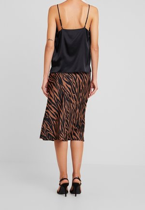 Femme portant un haut noir en satin à fines bretelles, une jupe midi marron et noire imprimé tigre, et des sandales noires à talons hauts, debout devant un fond gris.