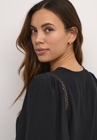 Blusa nera con maniche a sbuffo e dettagli in pizzo sulle spalle, texture liscia e silhouette rilassata. Design semplice ed elegante.