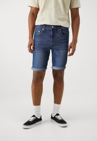 Jeansshorts i mörkblått med normal passform, uppvikt nederkant, fem fickor och en klassisk knappstängning. Stylad med svarta skor och vita strumpor.