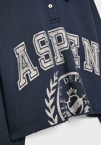Tamno plava majica s kragnom sa natpisom "ASPEN" u istrošenoj bijeloj boji, s dekorativnim amblemom s lovorovim listovima. Materijal izgleda kao mekani pamuk sa dodatkom.