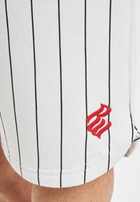 Shorts en tissu blanc avec des rayures verticales noires, ornés d'un logo rouge brodé à l'ourlet. Texture douce et coupe décontractée.