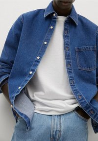 Blauwe denim shirt met een knoopsluiting en een borstzak, gedragen over een wit T-shirt. Lichtblauwe jeans zichtbaar bij de taille.