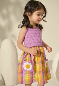Haut en tricot violet avec des bords festonnés ; jupe à carreaux colorée en jaune, rose et vert ; comporte deux poches avec des accents de fleurs blanches et jaunes.