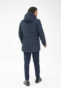 Veste bleu marine avec capuche, à coupe longue et fermeture éclair. Portée avec un pantalon bleu marine assorti et des chaussures noires.