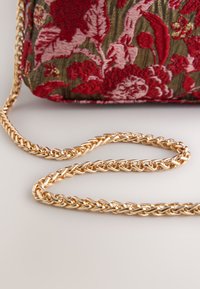 Goldfarbene Kettenriemen liegen auf einer Oberfläche, im Hintergrund ist eine Stofftasche mit roten und pinken Blumenstickereien zu sehen.