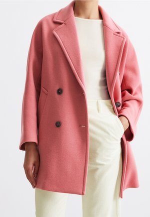 Personne portant un manteau croisé rose sur un haut blanc côtelé et un pantalon crème clair taille haute, main dans la poche.