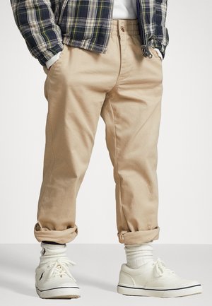 Una persona che indossa pantaloni color beige risvoltati, calzini bianchi, sneakers bianche con logo e una giacca a quadri con le mani nelle tasche.