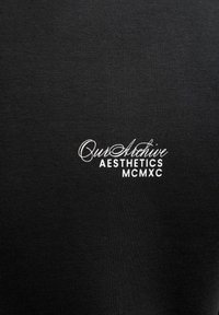 Mustast puuvillasest T-särgist, millel on valge tikandiga tekst: "Our Archive Aesthetics MCMXC", sileda soonilise tekstuuriga.