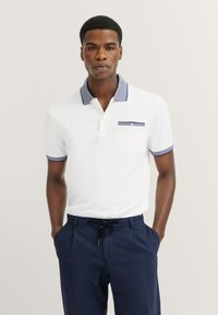Witte polo met een blauwe contrastkraag, korte mouwen en een borstzak met een blauwe rand; gecombineerd met marineblauwe broek.