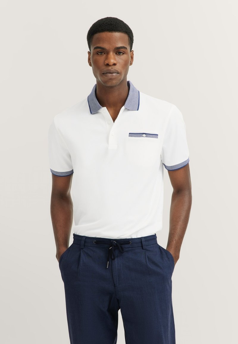 Witte polo met een blauwe contrastkraag, korte mouwen en een borstzak met een blauwe rand; gecombineerd met marineblauwe broek.