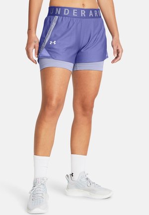 Donna che indossa pantaloncini sportivi a strati viola Under Armour, calzini bianchi e scarpe da corsa grigio chiaro, in piedi su sfondo bianco.