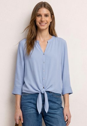 Jonge vrouw met lang bruin haar, een lichtblauw bloesje met knopen en gebonden in de taille, en een blauwe spijkerbroek, glimlachend tegen een bleke muur.