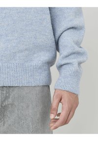 Pull en maille bleu clair avec des poignets et un ourlet côtelés, présentant une texture douce. Un pantalon gris est partiellement visible.