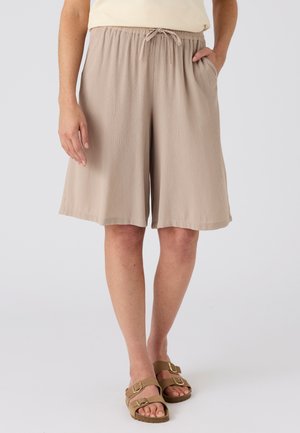 Frau trägt beige knielange, lockere Shorts mit elastischem Bund und Kordelzug, kombiniert mit beigen Sandalen mit doppeltem Riemen.