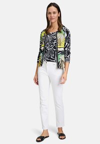 Multikleurige cardigan met abstracte patronen, over een zwart-wit grafisch T-shirt, gecombineerd met witte jeans en zwarte sandalen.