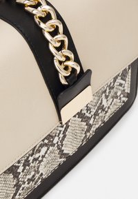 Sac à main structuré en cuir beige lisse, avec des accents noirs et un motif texturé imitation peau de serpent. Détail en chaîne dorée.