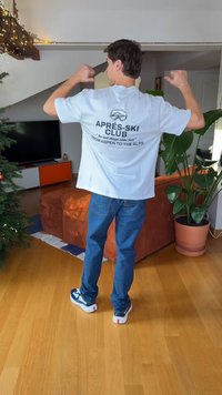 Witte T-shirt met zwarte tekst waarop staat "APRES-SKI CLUB" en "VAN ASPEN TOT DE ALPEN." Draagt blauwe jeans en casual sneakers.