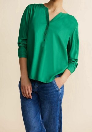 Persona vestida con una blusa verde de manga larga con botones y jeans azules, de pie con una mano en el bolsillo y la otra relajada a un lado.