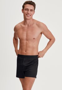 Zwarte katoenen boxershorts met een sluiting aan de voorkant en een elastische tailleband. Glad textuur en nauwsluitend ontwerp, model poseert tegen een neutrale achtergrond.