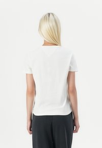Camiseta blanca de algodón con mangas cortas, que presenta un ligero corte en forma de V en la parte posterior, combinada con pantalones gris oscuro. La textura parece suave.