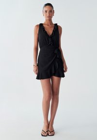 Schwarzes Kleid mit gerafftem V-Ausschnitt, Wickeldesign und Volant-Saum. Hergestellt aus glattem Stoff. Das Modell trägt dünne schwarze Sandalen.