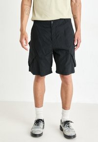 Shorts de cargo noirs en tissu texturé, avec une fermeture à bouton et des poches inclinées. Portés avec un t-shirt de couleur claire et des chaussures contrastées.