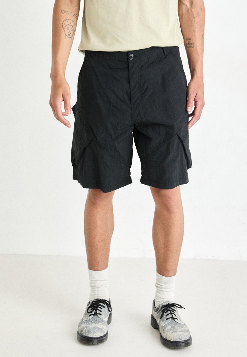 Shorts de cargo noirs en tissu texturé, avec une fermeture à bouton et des poches inclinées. Portés avec un t-shirt de couleur claire et des chaussures contrastées.