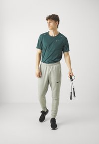 Zelena športna majica s kratkimi rokavi; sive trenirke; črne teniske; v roki drži vrvico za skakanje; logo Nike na majici in hlačah.