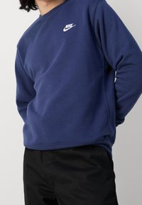 Tmavě modrá mikina z bavlněné směsi, s kulatým výstřihem a bílým logem Nike na levé straně hrudi. Pod ní jsou nošeny černé kalhoty.