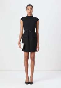 Robe noire sans manches avec un col haut, une silhouette ajustée, un détail de ceinture et deux poches à l'avant. Le modèle tient une petite pochette blanche.