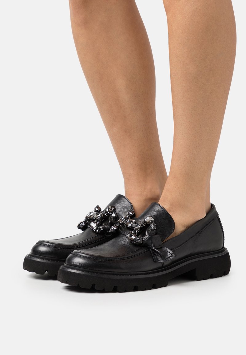 Mocassins en cuir noir avec des semelles épaisses portés pieds nus, chacun décoré de grandes pierres précieuses sombres et brillantes sur le dessus.