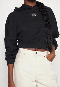 Czarna krótka bluza z kapturem o prążkowanej fakturze i naszywką z logo z przodu, zestawiona z wysokim stanem kremowymi dżinsami z zapięciem na guziki i kieszeniami.