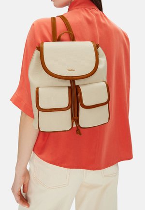 Žena v korálovém tričku a krémovém batohu s hnědým lemováním a dvěma předními kapsami, zachycená zezadu.