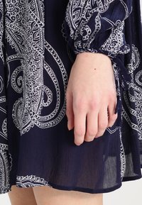 Marineblaues Kleid mit weißen Paisleymustern, langen Ärmeln mit elastischen Bündchen und einem fließenden, strukturierten Stoff. Die Hand ruht auf dem Stoffsaum.