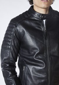 Chaqueta de cuero negra con cierre de cremallera, acentos acanalados en los hombros y un cuello alzado con botones a presión. Acabado texturizado.